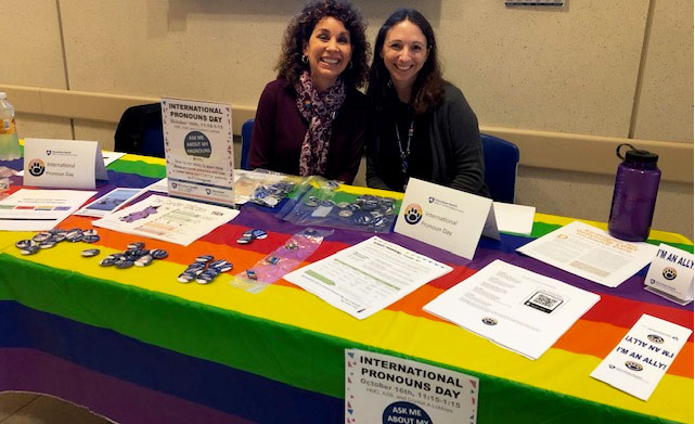 Two smiling people are pictured sitting at a table covered in a rainbow tablecloth that has handouts, buttons and other information.