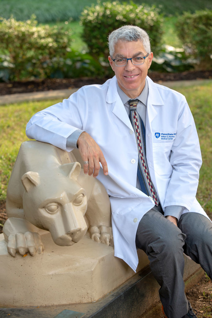 A man in a medical coat sits with a statue of a lion in an outdoor garden, smiling professionally.