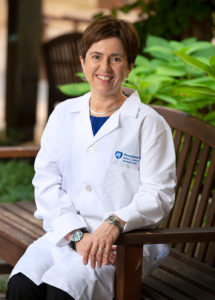 A woman in a medical coat sits on a bench in an outdoor garden, smiling professionally.