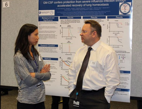 A man stands in front of an academic poster with his hands in his pockets, talking with a woman.