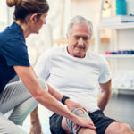 A female physical therapist kneels down as she places her hands on both sides of a man's knee. He is seated on the floor, with a brace on the knee. Various physical therapy equipment is in the background, out of focus.