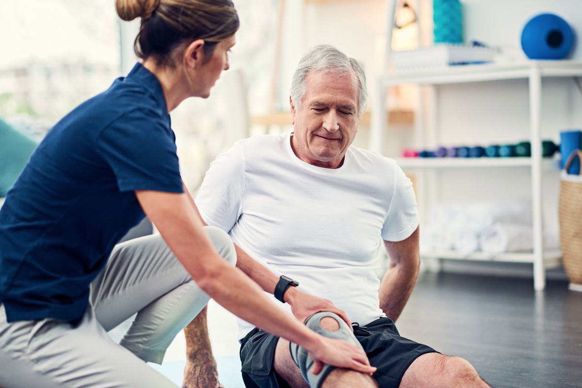 A female physical therapist kneels down as she places her hands on both sides of a man's knee. He is seated on the floor, with a brace on the knee. Various physical therapy equipment is in the background, out of focus.