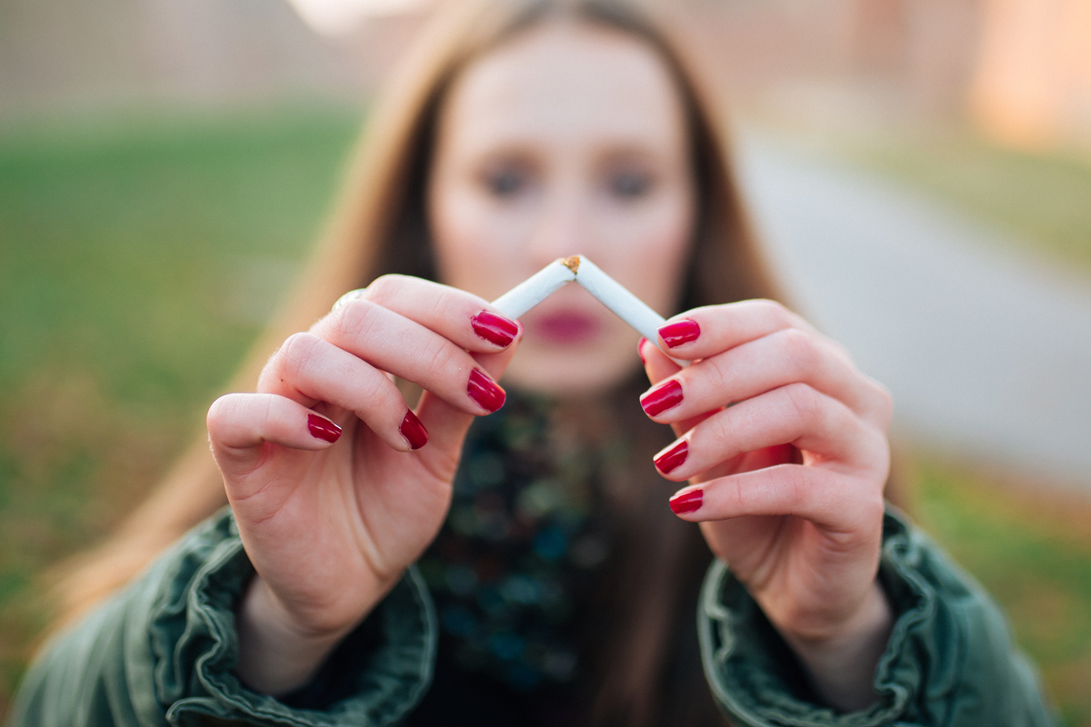 A woman stands outdors, facing the camera. She holds up a cigarette as she breaks it in half in front of the camera.