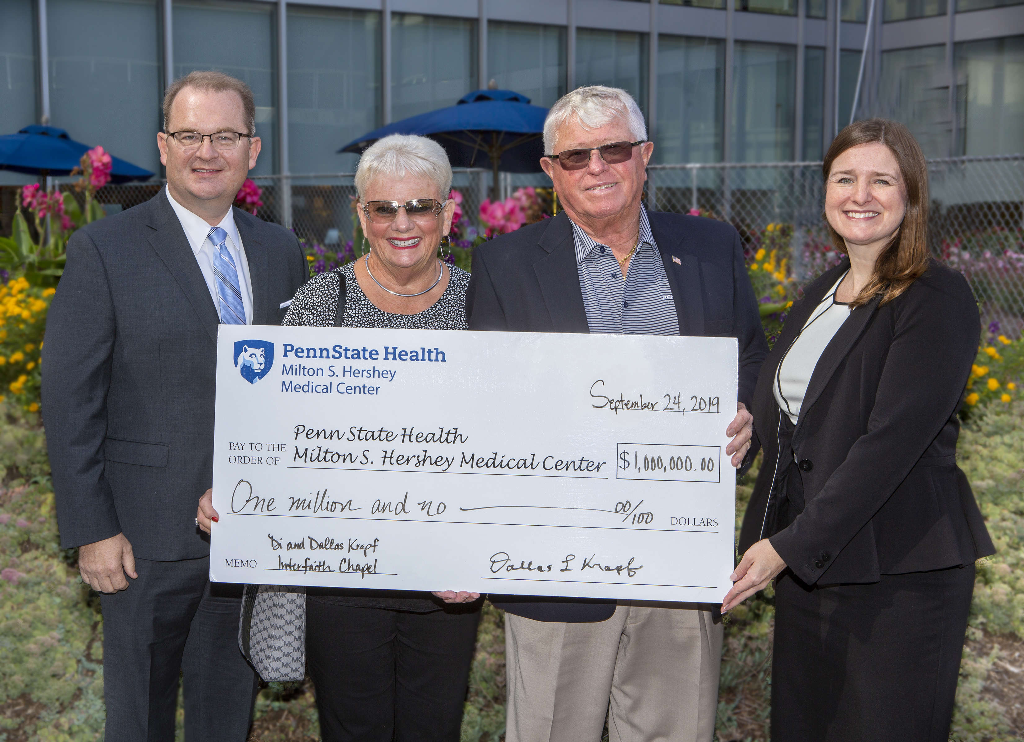 Four people – two men and two women – pose outside for a photo holding an oversized check made out to Penn State Health Milton S. Hershey Medical Center, in the amount of $1 million. It’s signed by Dallas Krapf. A building with windows is in the background, as are bushes and flowers.