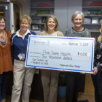 Five women pose with an oversized check in the amount of $10,000, made out from Highmark to Penn State Health. The Memo line reads: Eye glasses for students in need. A desk and office shelves are in the background.