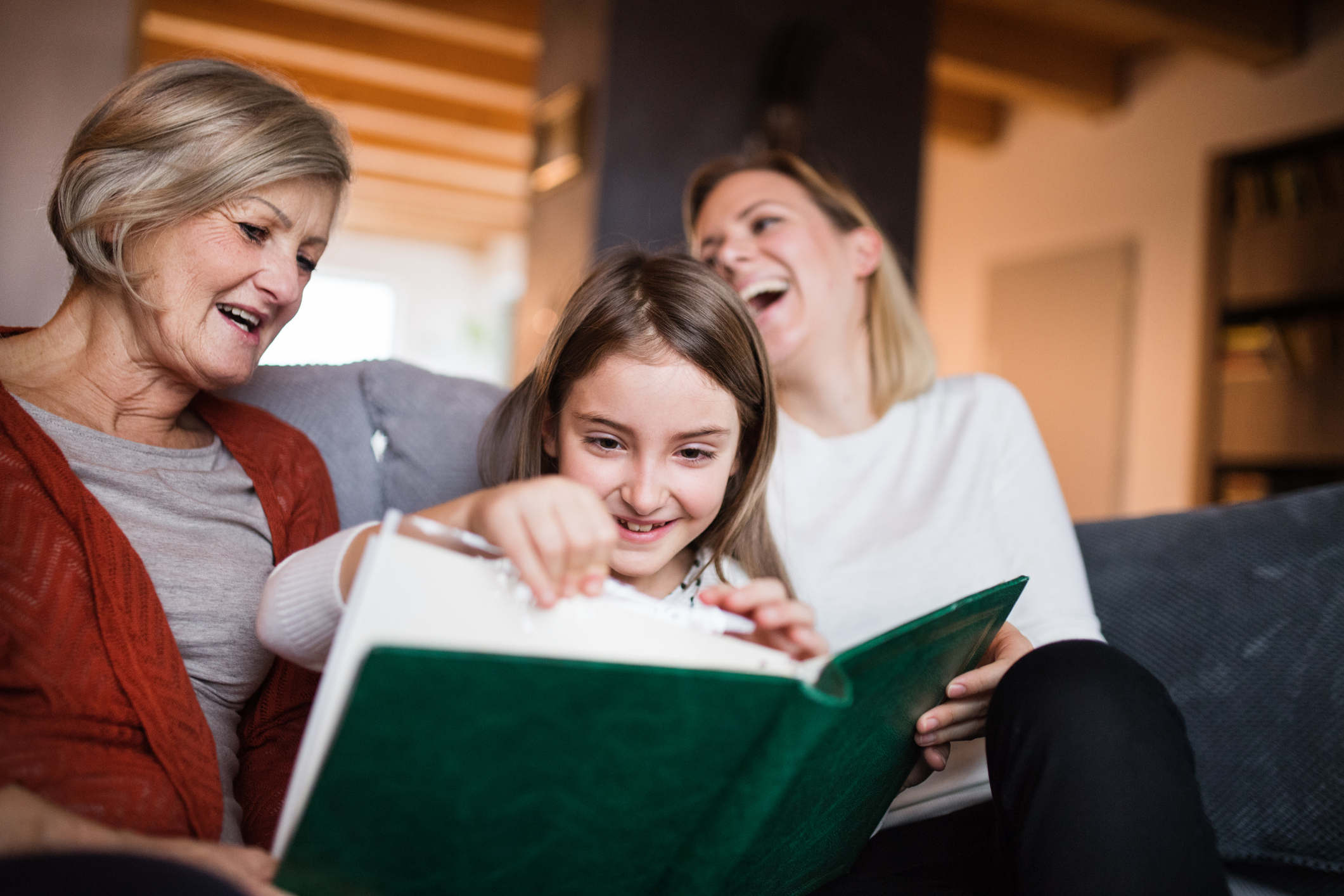 A young girl sits between two women on a couch. She holds and pages through a large photo album as the women look on, smiling. A bookshelf and window are in the background.