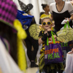 Trinity Stryker hoists a decorative bag. She’s wearing sunglasses, wings and antennae. All around her are other children and adults in costume holding bags.