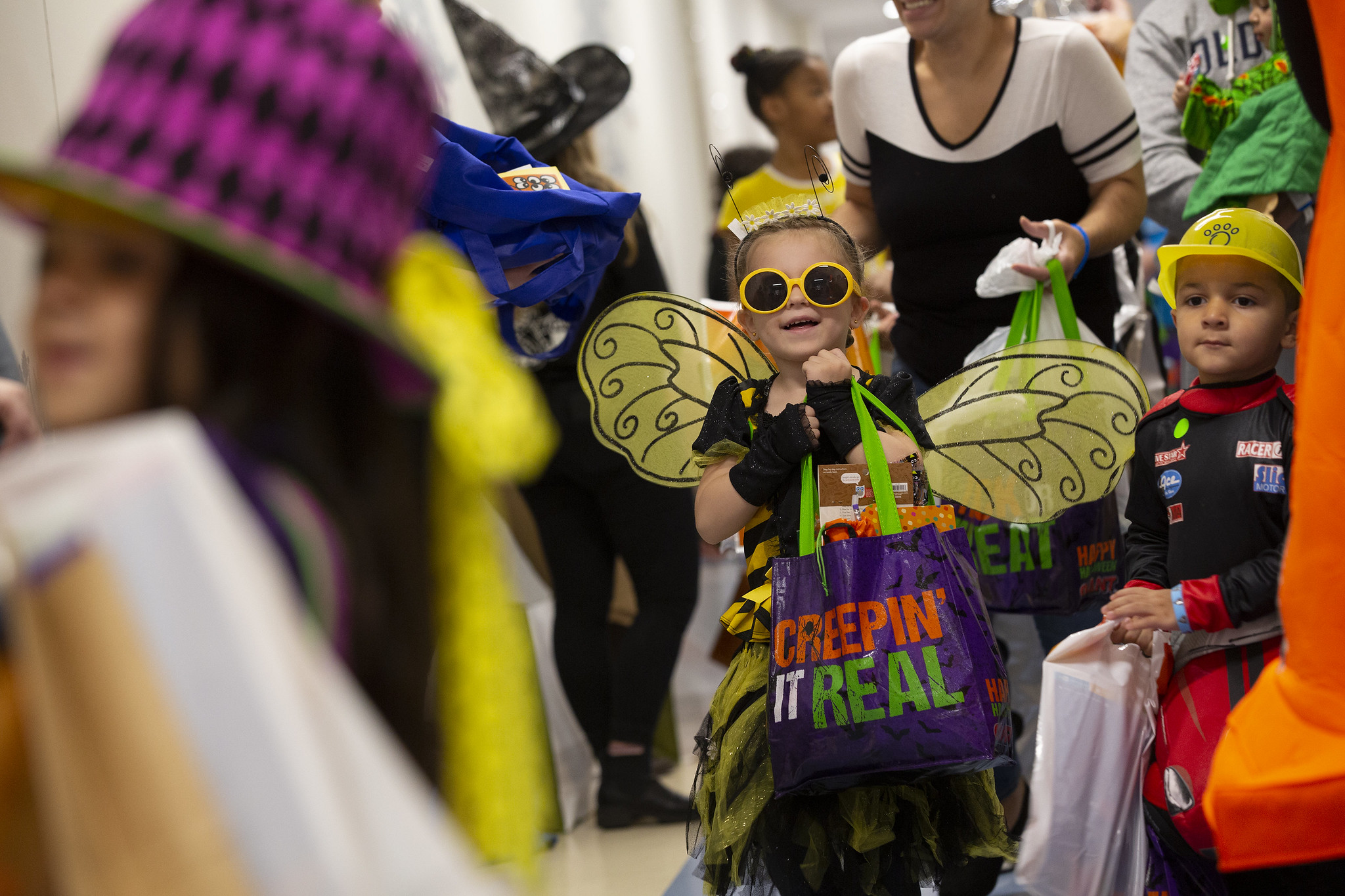 Trinity Stryker hoists a decorative bag. She’s wearing sunglasses, wings and antennae. All around her are other children and adults in costume holding bags.