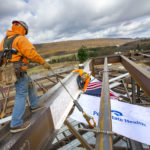 Two construction workers guide a steel beam into place on the top of a steel frame structure