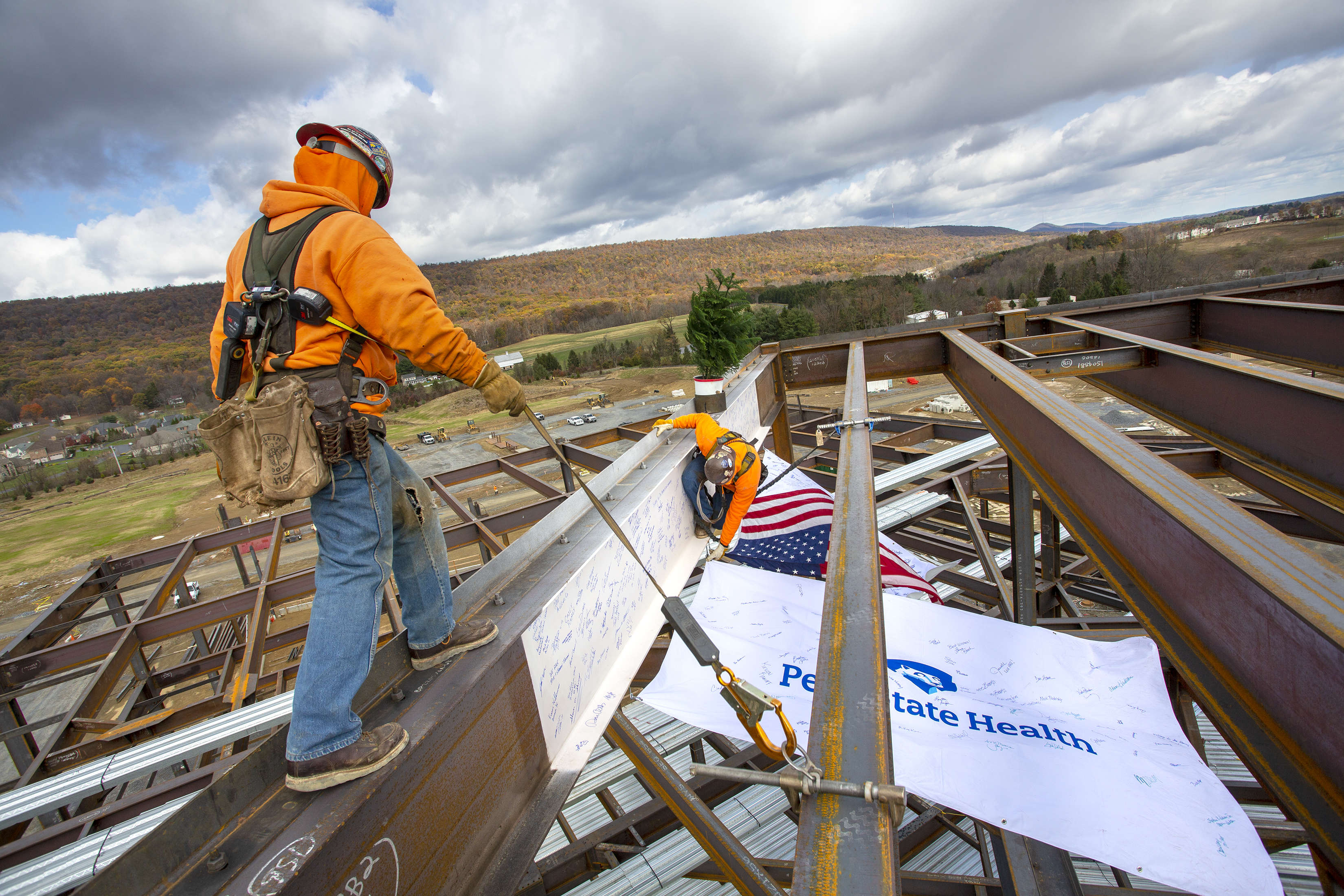 Two construction workers guide a steel beam into place on the top of a steel frame structure