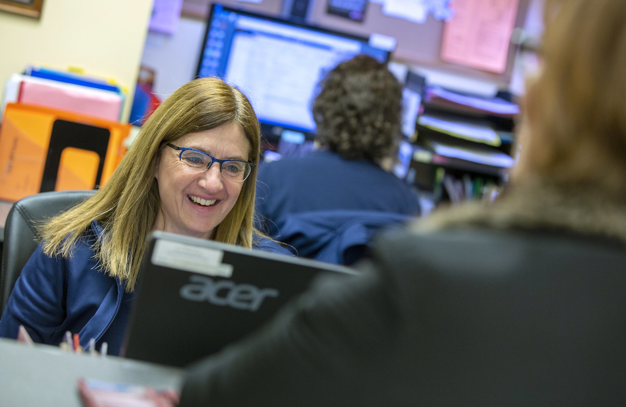 Medical office associate Laura Bjanes smiles as she looks at her computer monitor. She has shoulder-length hair, glasses and is wearing a polo shirt. In front of her a woman is seeing from behind standing at the counter. Another woman employee is behind Laura working at a computer.