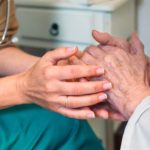 Two pairs of hands intertwine. One pair belongs to someone wearing a stethoscope and scrubs. The other belongs to someone in a white gown.