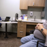 Kat Schoenknecht sits at a desk in a doctor’s office with a laptop opened. She holds a sheet of paper and smiles at a man seated across from, who sits with his hands folded on his stomach.