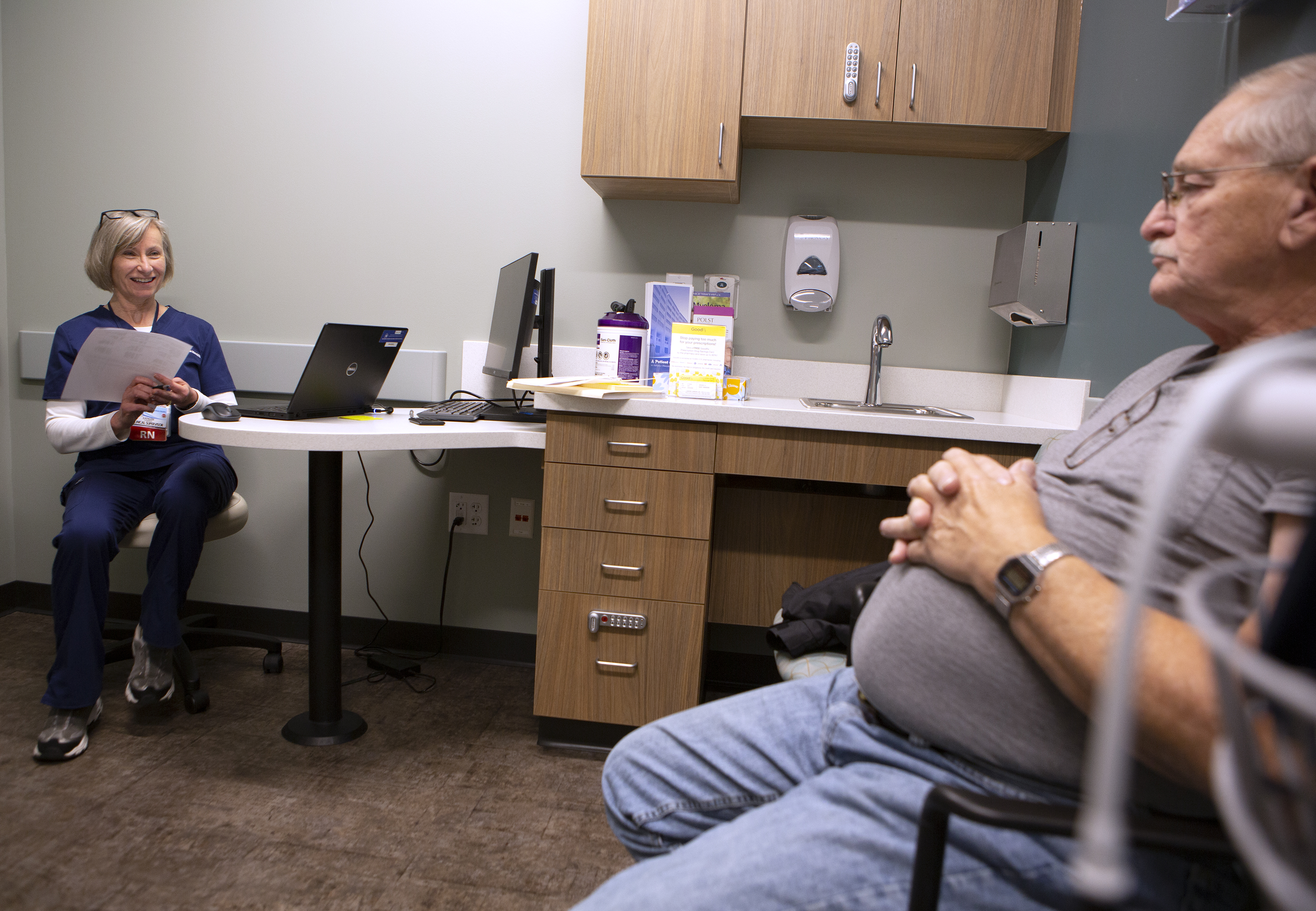 Kat Schoenknecht sits at a desk in a doctor’s office with a laptop opened. She holds a sheet of paper and smiles at a man seated across from, who sits with his hands folded on his stomach.