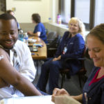 Dr. Cameron Gilliard smiles and exposes his arm. A bandage is affixed to his shoulder. Seated across from him, Registered Nurse Monica Hardy, who has just administered a flu shot, smiles.