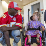 LeSean McCoy, wearing a Kansas City Chiefs uniform and hat, kneels down to talk with a young girl in a wheelchair. She looks back at him.