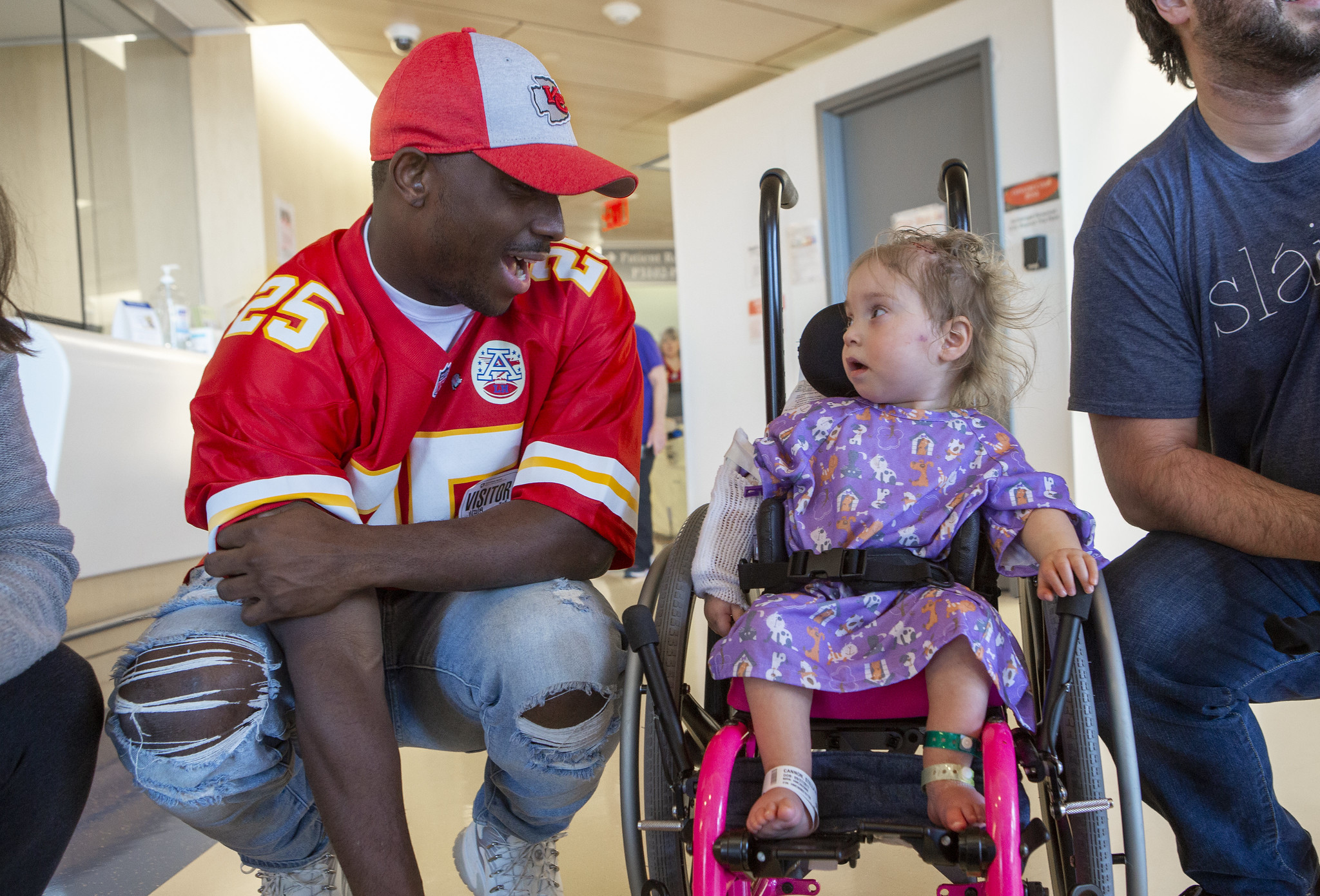 LeSean McCoy, wearing a Kansas City Chiefs uniform and hat, kneels down to talk with a young girl in a wheelchair. She looks back at him.