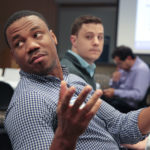 A student is seen looking over his shoulder and gesturing with his hand. Another student is visible in the background, watching the first one.