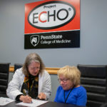 Two women sit at a table working with a Project ECHO logo on the wall behind them.