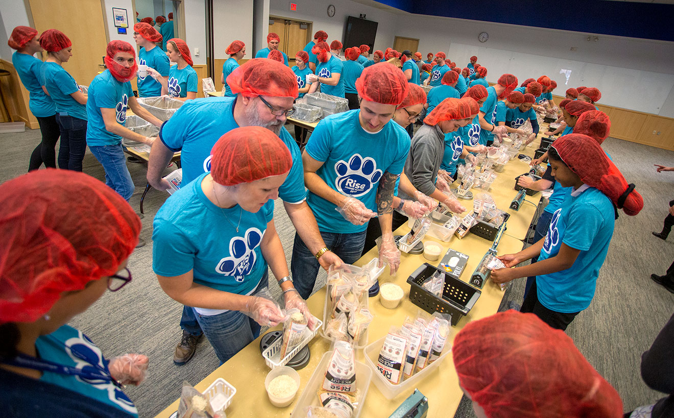 A group of students wearing matching T-shirts and hairnets is seen packing food items in a large room.