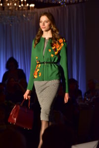 A model walks the runway at the 2019 Neiman Marcus Runway Show. She wears a checkered dress, a flowered sweater over it, and carries a handbag in her right hand.