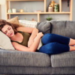 A woman wearing a tank top and jeans lays on her side on a sofa, holding onto a small pillow with two hands over her stomach. A shelf with various household items is in the background.