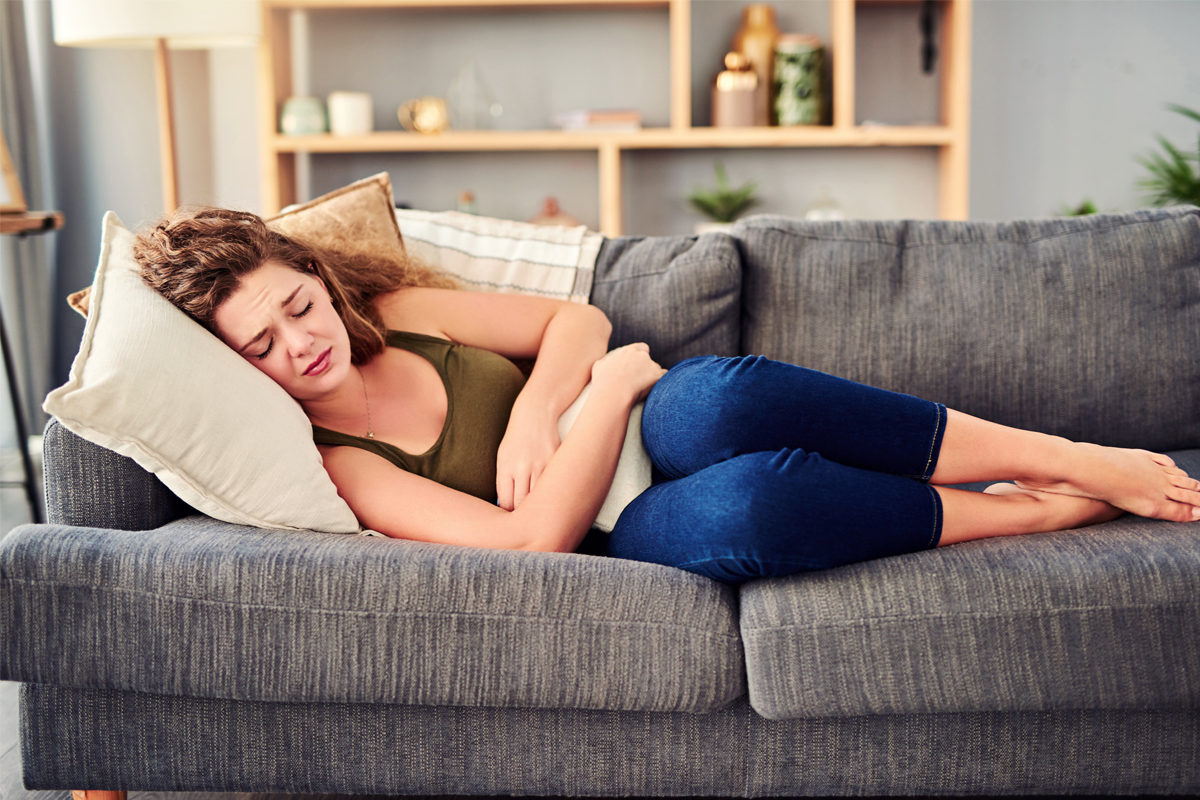 A woman wearing a tank top and jeans lays on her side on a sofa, holding onto a small pillow with two hands over her stomach. A shelf with various household items is in the background.