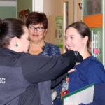 Steph Reed stands in front of Gabby Wallace and attaches something to her shirt while Judy Himes and Christine Ewing look on.