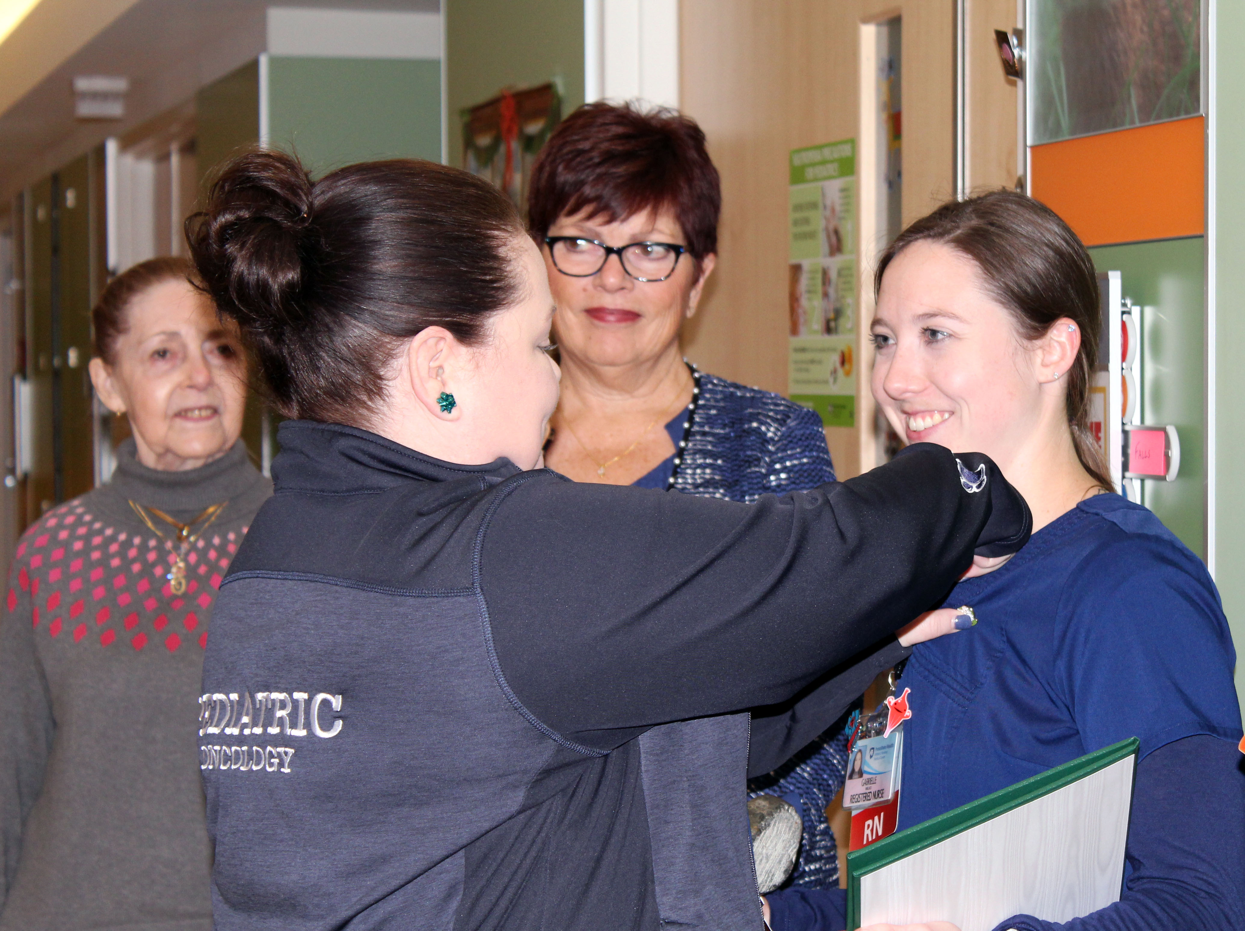 Steph Reed stands in front of Gabby Wallace and attaches something to her shirt while Judy Himes and Christine Ewing look on.