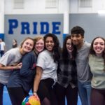 Six high school students – five female and one male – pose for a group photo with arms around each other. They’re in a school gym, where a wall with the word “PRIDE” is in the background.