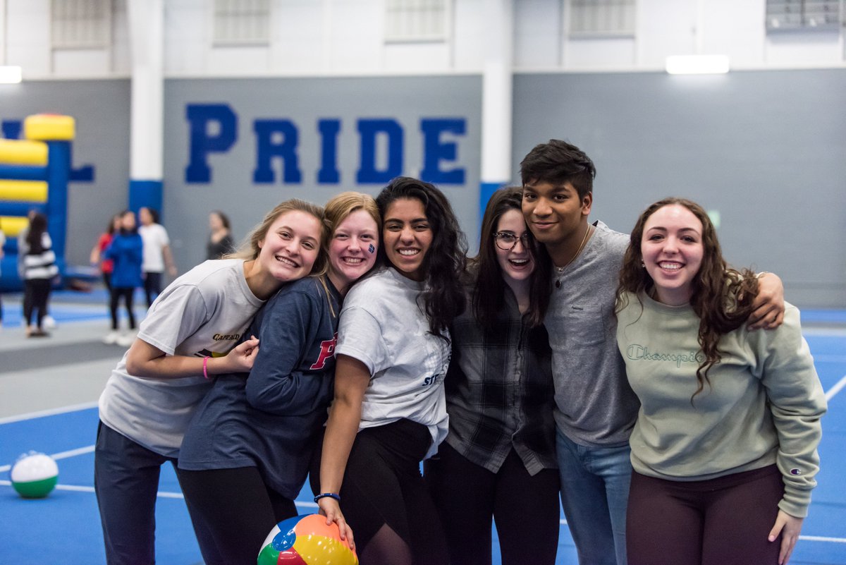 Six high school students – five female and one male – pose for a group photo with arms around each other. They’re in a school gym, where a wall with the word “PRIDE” is in the background.