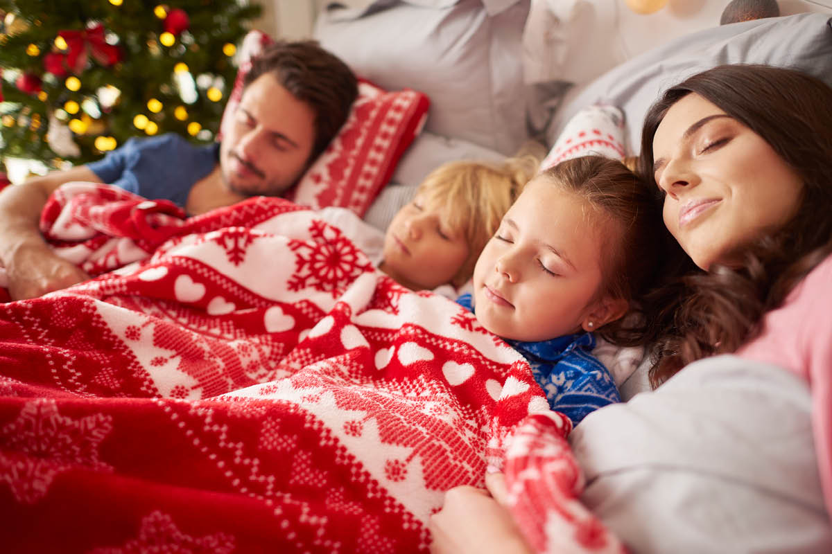 A man, woman and two young children lay down, sleeping, covered in a holiday-themed blanket. A lit Christmas tree is in the background.