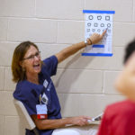 Julie Groh smiles as she points to a shape on an eye chart. She has shoulder-length hair and is wearing glasses, scrubs and an ID badge with “RN” on it. Two children are in the foreground out of focus.