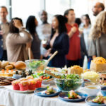 A table with plates and bowls of food is in the foreground. It includes a salad, cookies, muffins, bread and various hors d’oeuvres. In the background, several people are standing and talking, some of them holding drinks.