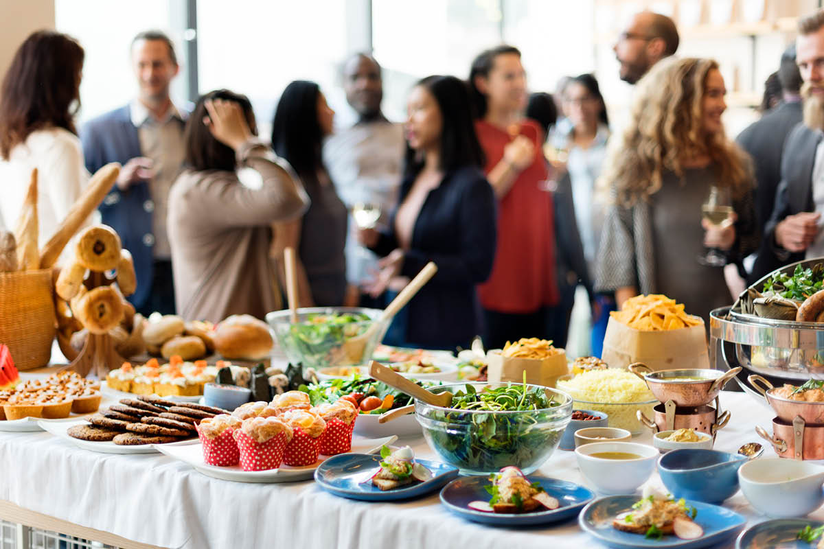 A table with plates and bowls of food is in the foreground. It includes a salad, cookies, muffins, bread and various hors d’oeuvres. In the background, several people are standing and talking, some of them holding drinks.