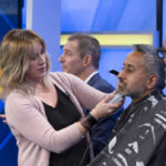A man sits on a television show stage, having his beard shaved.