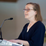 A woman stands at a lectern, speaking into a microphone. She is wearing glasses and looking intently at the audience.