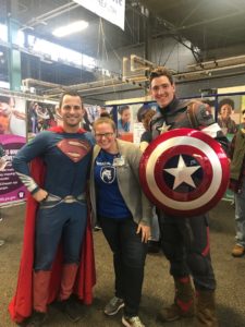 A woman smiles with two men dressed up as superheros