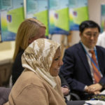 Dr. Alawia Suliman speaks at a table while others look on. On the table are half eaten lunches and water bottles. Behind them, out of focus, are posters on easels.