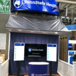 The Penn State Health front porch is a wooden stage with a porch with shingles rising behind it, lined by a railing.