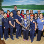 A cluster of people pose and smile for a camera. They stand on a hardwood floor in front of a mural depicting scenery.