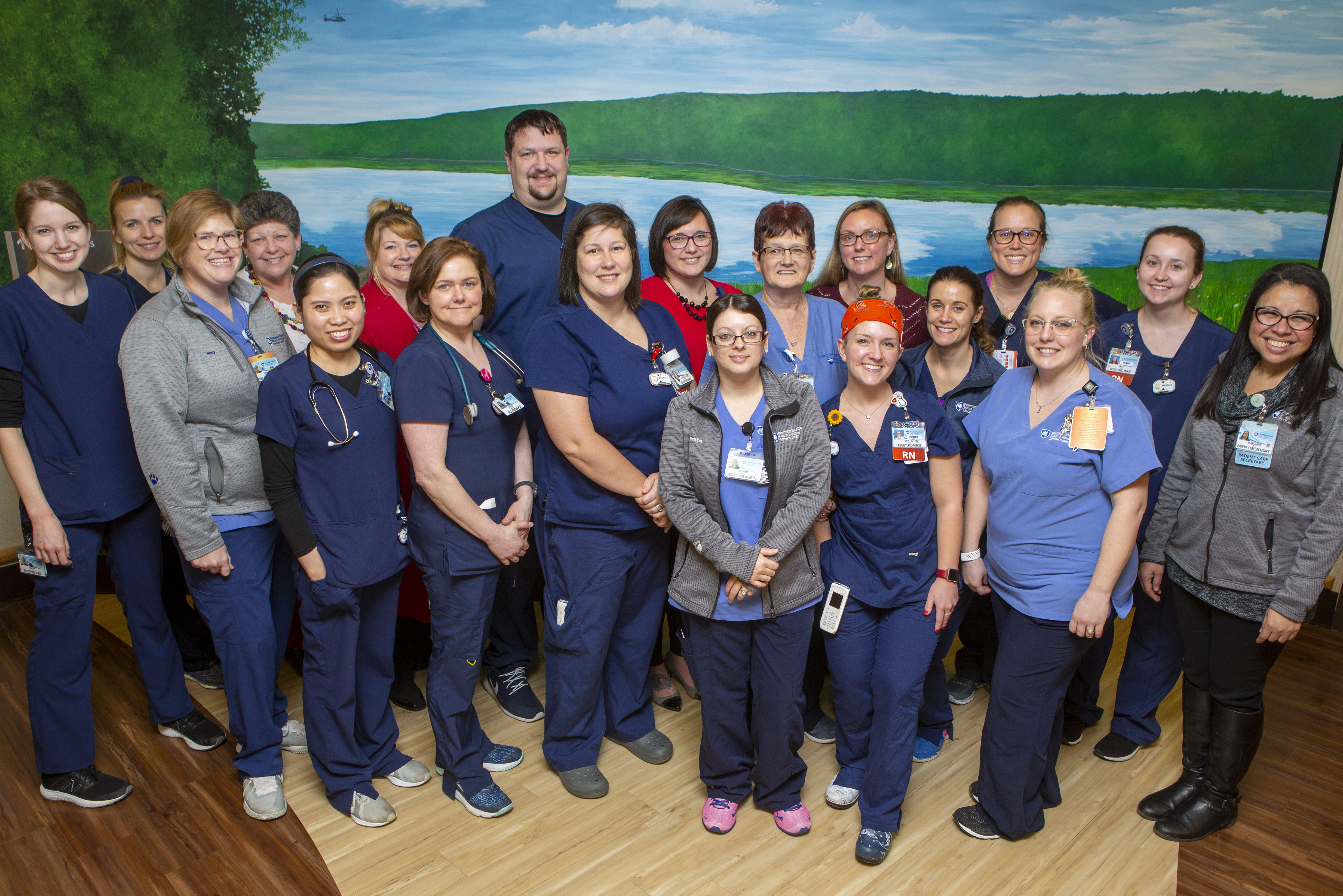 A cluster of people pose and smile for a camera. They stand on a hardwood floor in front of a mural depicting scenery.