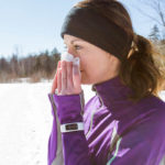 A woman stands outside in winter, wearing workout clothes including a jacket and headband. She holds a tissue up to her nose. Snow is on the ground and trees are in the background.