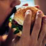 A close-up of a man with a short beard and mustache holding a burger on a bun, about to take a bite out of it. He holds the burger with both hands.