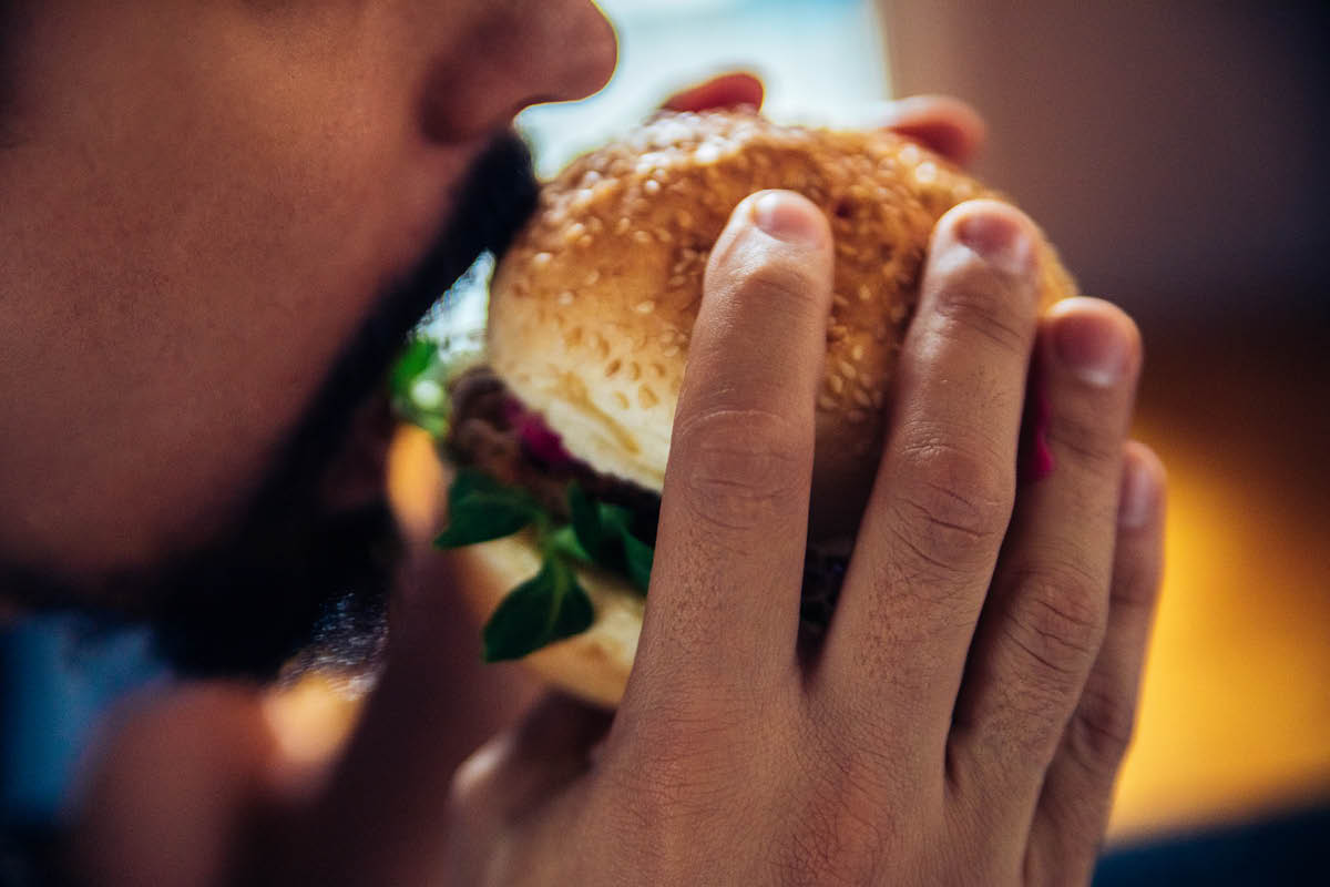 A close-up of a man with a short beard and mustache holding a burger on a bun, about to take a bite out of it. He holds the burger with both hands.