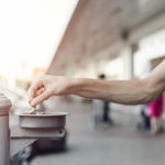 An outstretched hand holds a cigarette butt as its remnants are snuffed in an ashtray, which is mounted to a pole outdoors. Vehicles and a building are in the background, slightly out of focus.