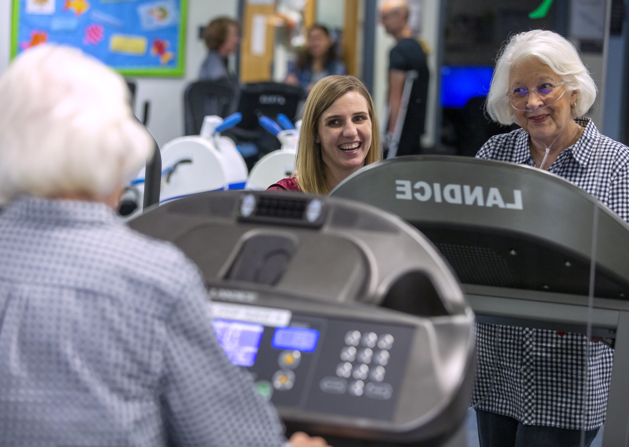A young woman smiles as an older woman wearing a nasal cannula and a checkered dress and standing on a treadmill. They are in the Health and Wellness Center on the Hershey Medical Center campus. Three people are standing behind them out of focus.