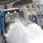 A plow flings a pile of snow into the air. Behind it, someone steps out of an ambulance.