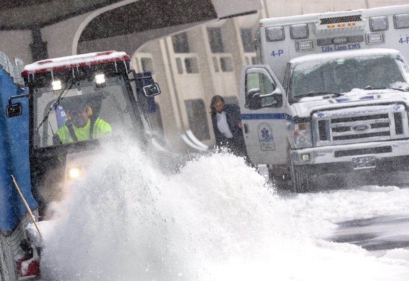 A plow flings a pile of snow into the air. Behind it, someone steps out of an ambulance.