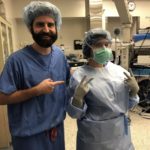 Dr. Eric Pauli poses with registered nurse Kiersten Marie in the operating room. Marie wears a surgical gown, gloves, mask, hair net and glasses. Pauli, who has a beard and moustache, wears scrubs and points his index finger toward Marie. Operating equipment is in the background.